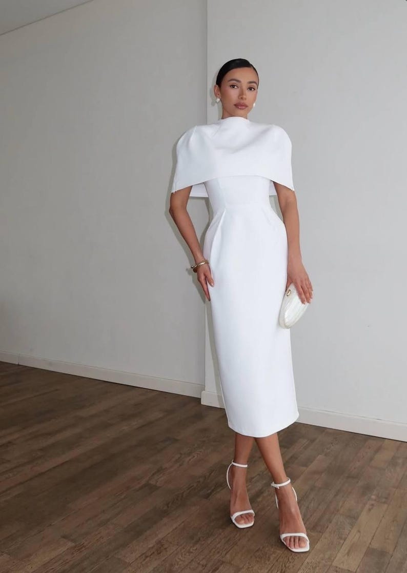 K&ouml;nnte beinhalten: Ein wei&szlig;es Midikleid mit einem einzigartigen, kapuzenartigen &Uuml;berzug an den Schultern. Das Kleid hat eine figurbetonte Silhouette, einen hohen Ausschnitt und wird mit wei&szlig;en Riemchen-Heels und einer Clutch kombiniert. Der Look ist elegant und anspruchsvoll.