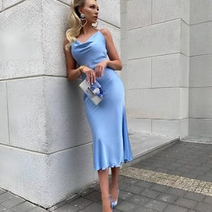 Puede incluir: Vestido lencero de sat&eacute;n azul claro con cuello de cisne y largo midi. El vestido se combina con zapatos de tac&oacute;n a juego con punta y un peque&ntilde;o clutch. La mujer lleva pendientes en forma de coraz&oacute;n y una pulsera.
