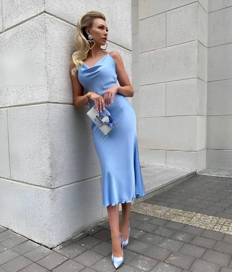 Puede incluir: Vestido lencero de sat&eacute;n azul claro con cuello de cisne y largo midi. El vestido se combina con zapatos de tac&oacute;n a juego con punta y un peque&ntilde;o clutch. La mujer lleva pendientes en forma de coraz&oacute;n y una pulsera.