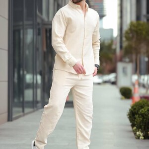 May include: A light beige linen shirt and matching trousers set. The long-sleeved shirt is buttoned and paired with straight-leg trousers. The outfit is completed with white trainers. The person is wearing sunglasses.