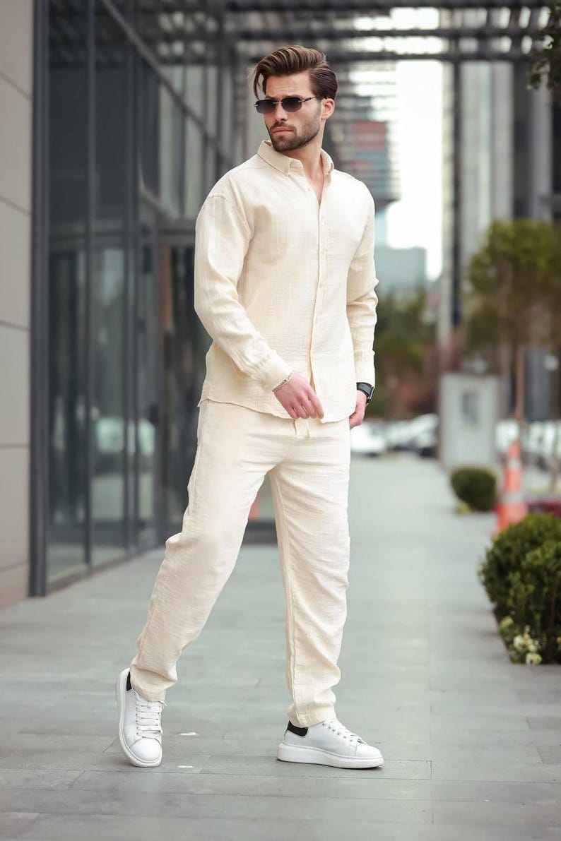 May include: A light beige linen shirt and matching trousers set. The long-sleeved shirt is buttoned and paired with straight-leg trousers. The outfit is completed with white trainers. The person is wearing sunglasses.