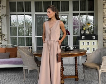 May include: A sleeveless, light pink jumpsuit with a tie-up top and wide-leg pants. The outfit includes a small brown crossbody bag. The setting is an outdoor patio with a stone wall and large windows.