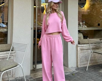 May include: A light pink sweatshirt and matching wide-leg sweatpants set. The outfit includes a cropped crew neck sweatshirt and high-waisted pants. The model is wearing a light pink baseball cap and white sneakers. The outfit is made of a soft, comfortable material.