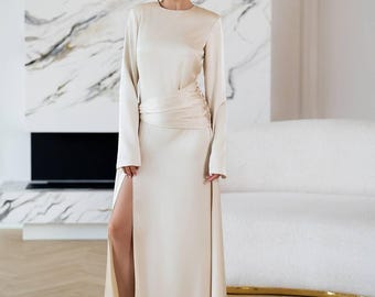 May include: A floor-length champagne-colored satin gown featuring long sleeves, a round neckline, and a high side slit. The waist is accented with a gathered, wrapped fabric detail that creates a draped effect across the midsection.