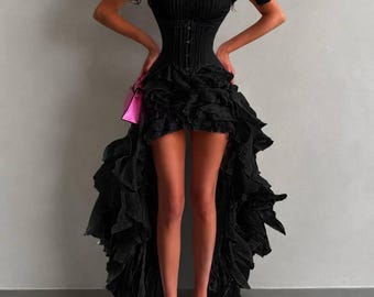 May include: A black off-the-shoulder dress featuring a corset-style bodice with vertical ribbing and a front button closure. The skirt has a high-low hemline with voluminous, layered ruffles that extend to the floor at the back and sides.