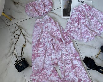 May include: A matching set featuring a strapless tube top, wide-leg trousers, and a pair of shorts, all crafted from white fabric with a pink floral toile pattern. The set is displayed alongside a black quilted handbag with a gold chain strap.