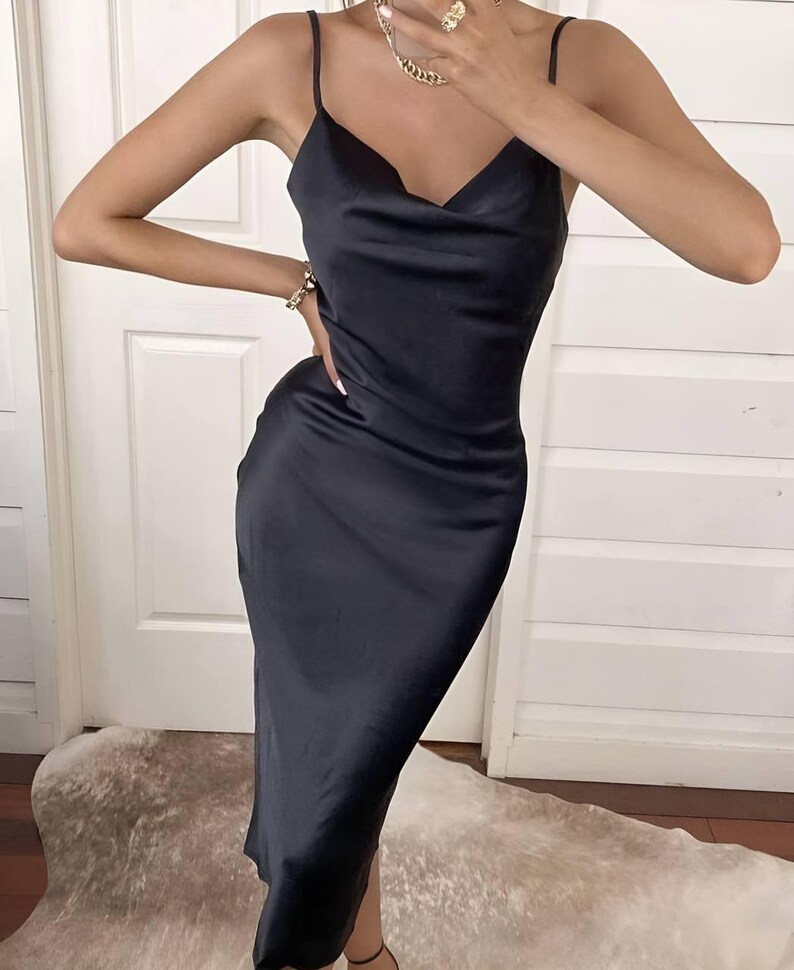 Puede incluir: Vestido midi negro de sat&eacute;n con cuello drapeado y tirantes finos. El vestido tiene una silueta elegante y entallada. Se ven un collar y una pulsera dorados. El vestido es adecuado para eventos formales.