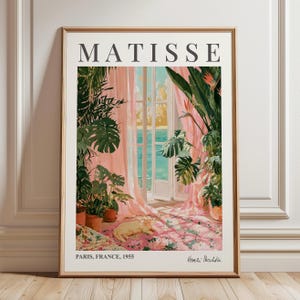 May include: A framed art print titled MATISSE featuring a scene of a sunlit room with large green potted plants, sheer pink curtains, and a view of the sea. A light-coloured cat rests on a floral rug. Text reads PARIS, FRANCE, 1955 and Henri Matisse.