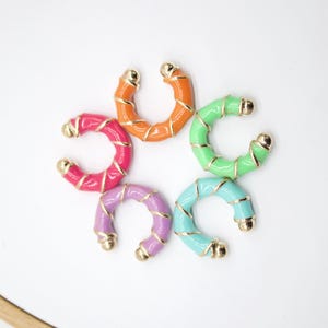 May include: A collection of six colorful, C-shaped earrings. The earrings are in shades of pink, orange, green, blue, and purple, with gold-colored accents. The earrings are arranged in a circular pattern against a white background.