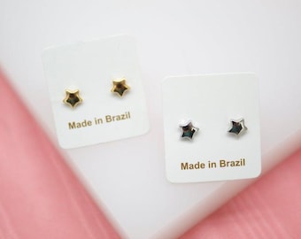 Puede incluir: Dos pares de pendientes de estrella. Un par es dorado, el otro plateado. Cada par se muestra en una tarjeta blanca con el texto "Made in Brazil". El fondo es blanco y rosa.