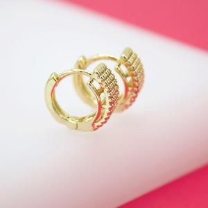 May include: A pair of gold-coloured hoop earrings. The earrings feature a textured design on the outer edge, with small, closely spaced details. The hoops are clasped shut. The background is a soft white and pink.