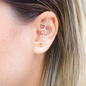 Puede incluir: Puño de oreja dorado con tres estrellas. El puño de oreja está diseñado para usarse a lo largo de la oreja externa. La joya tiene una estética delicada y celestial. La persona de la imagen tiene el pelo rubio.