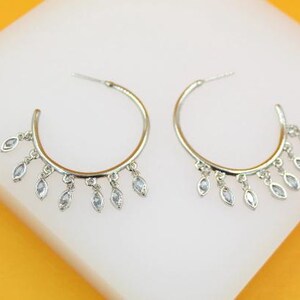 May include: A pair of silver-toned hoop earrings. Each earring features a half-circle design with dangling, teardrop-shaped, clear gemstones. The earrings are displayed on a white surface with a yellow background.