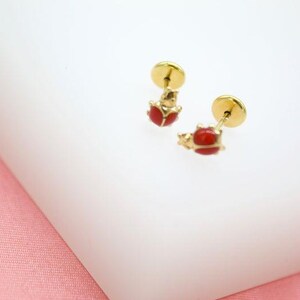 May include: A pair of gold-colored stud earrings featuring a ladybug design. The ladybugs have red bodies and are set against a white background. The earrings are small and delicate, with a post and backing.