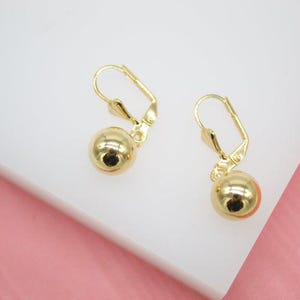 Puede incluir: Un par de pendientes colgantes dorados. Cada pendiente presenta una pequeña esfera redonda y pulida suspendida de un cierre de palanca. Los pendientes se muestran sobre un fondo blanco y rosa.