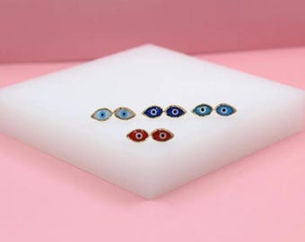 May include: A collection of gold-toned stud earrings, each featuring an eye design. The eyes are in various colors, including blue, red, and teal, with a gold border. The earrings are displayed on a white surface against a pink backdrop.