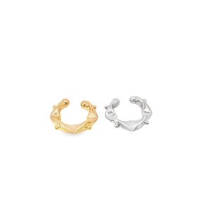 Puede incluir: Dos puños de oreja, uno dorado y otro plateado, se muestran sobre un fondo blanco. Cada puño presenta un diseño texturizado distintivo con un punto central y pequeñas protuberancias redondeadas. Los puños están diseñados para envolver la oreja.