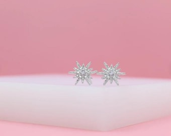 May include: Two silver starburst stud earrings, each with clear, sparkling details. The earrings are displayed on a white surface against a pink backdrop. These earrings, with their star-like design, would make a lovely gift or a stylish addition to any jewellery collection.