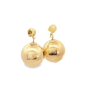 May include: A pair of gold-colored, spherical drop earrings. Each earring features a small, round stud connected to a larger, polished ball. The earrings have a reflective surface, and the design is simple and elegant.