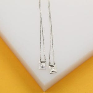 May include: Two silver-colored necklaces with small rectangular pendants. Each pendant features a religious image. The necklaces hang on a white surface with a yellow background. The chains are delicate and the pendants are detailed.