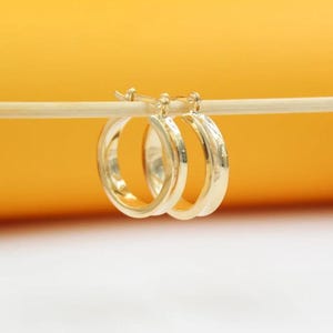 May include: A pair of gold-colored hoop earrings. The earrings are round and have a smooth, polished surface. They are displayed against a yellow and white background.