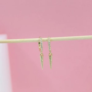 May include: A pair of gold-colored hoop earrings with small, clear stones along the hoop. Each earring has a gold-colored, cone-shaped charm hanging from it. The earrings are displayed against a light pink background.