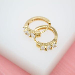 May include: A pair of gold-colored hoop earrings. Each earring features a row of clear, sparkling gemstones. The earrings are displayed on a white surface with a pink background, creating a contrast that highlights the jewelry's design.