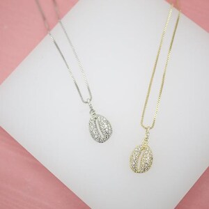 Peut inclure: Deux colliers avec des breloques pendantes. L'un a une chaîne argentée et un pendentif argenté incrusté de petites pierres transparentes. L'autre a une chaîne dorée et un pendentif doré avec des pierres transparentes. Les colliers sont présentés sur une surface blanche sur fond rose.