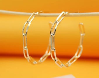 May include: A pair of gold-colored hoop earrings. Each earring features a chain link design, with elongated rectangular links forming a semi-circle. The earrings are displayed against a bright yellow background.