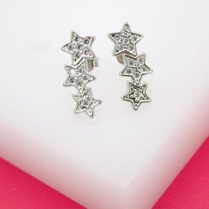 May include: A pair of silver-toned star earrings. Each earring features three graduated stars, each encrusted with small, clear crystals. The earrings are displayed on a white surface with a pink background.