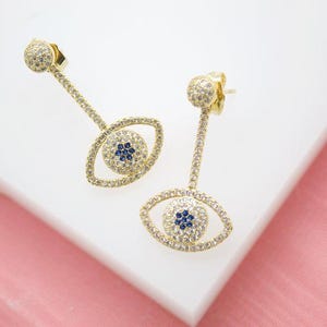 May include: A pair of gold-colored, dangling earrings in the shape of an eye. The earrings are encrusted with small, clear crystals and feature a blue crystal center. The earrings are displayed on a white and pink surface.