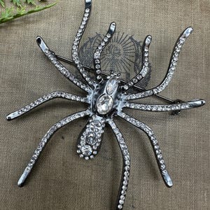 May include: A spider-shaped brooch crafted from dark metal, featuring eight long legs encrusted with small clear rhinestones. The body is adorned with larger oval and round clear crystals, creating a sparkling, detailed insect design.