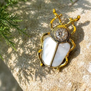 May include: A gold-toned beetle brooch with a white, pearlescent body and a sparkling crystal center. The brooch has detailed antennae and is set against a textured, neutral-toned surface, with a sprig of green foliage in the background.
