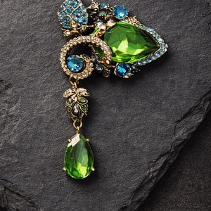 May include: An ornate brooch with a gold-tone setting, featuring a large pear-shaped green gemstone and smaller blue and green stones. The brooch has a detailed, floral-inspired design with a dangling green teardrop-shaped stone.