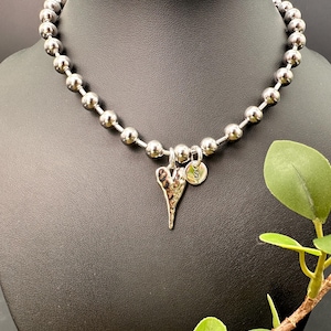 May include: Silver-toned beaded necklace with a heart pendant and a small circular charm with the letter "S". The necklace is displayed on a black mannequin. Green leaves are in the foreground.