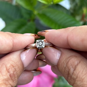 May include: A silver ring with a square, geometric design. The ring is held between two hands with manicured nails, set against a backdrop of pink flowers and green foliage. The ring's design is a series of intersecting lines within the square.