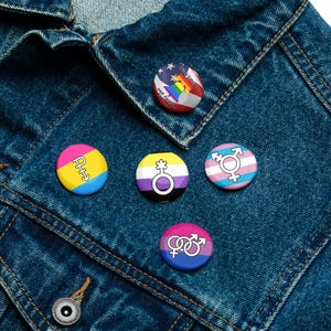 May include: A collection of round, colorful pins with various LGBTQ+ pride symbols and flags, including the American flag with a rainbow, and symbols for pansexual, non-binary, and transgender identities. The pins are displayed on a denim jacket.