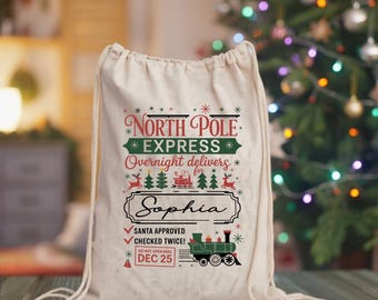 May include: A cream-colored drawstring bag with the text "North Pole Express" in red and green. The bag includes the name "Sophia" and a graphic of a green train. The text also says "Santa Approved" and "Dec 25".