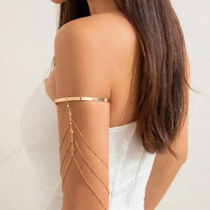 Peut inclure: Chaîne de bras dorée avec une bande fine en haut et plusieurs brins de chaîne avec de petites perles transparentes qui pendent.