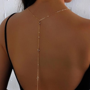Puede incluir: Un collar de cadena dorada con pequeñas piedras transparentes que cuelgan de él. El collar se lleva en la parte posterior del cuello y cuelga por la espalda.