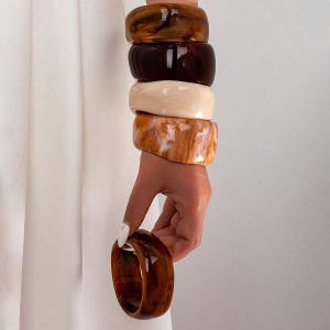 May include: A collection of chunky, colorful bangles in shades of brown, cream, and orange. The bracelets are stacked on a person's arm, with one held in the hand. The bracelets have a glossy finish and a marbled appearance, perfect for adding a pop of color.