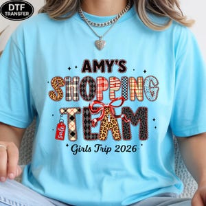May include: Light blue t-shirt with the text "AMY'S SHOPPING TEAM" in a mix of patterns and colors. The design includes a bow and the words "Girls Trip 2026". A small "Sale" tag hangs from the letter "T".