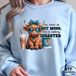 May include: Light blue sweatshirt featuring a cartoon highland cow wearing sunglasses and a bow, with the text "I'm not a HOT MESS, I'm a spicy DISASTER." The graphic includes a coffee drink and a blue boot.