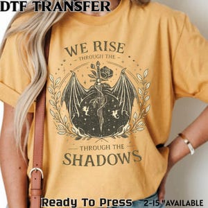May include: Mustard yellow t-shirt with the text "WE RISE THROUGH THE SHADOWS". The graphic features a sword, rose, and dragon wings. The words "Ready To Press" and size availability are also printed on the shirt.