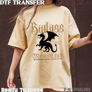 May include: A light yellow t-shirt with the words "Badass Moms Club" and a black dragon graphic. The text "Motherhood Hasn't Killed Us Yet" is below the graphic. The shirt also has the text "Ready To Press" and "2-15 AVAILABLE".