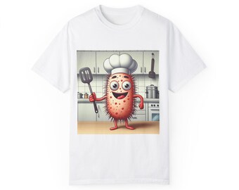 Op de afbeelding: Wit T-shirt met een cartoonillustratie van een rood en wit micro-organisme dat een koksmuts draagt en een spatel vasthoudt. Het micro-organisme bevindt zich in een keukensituatie.