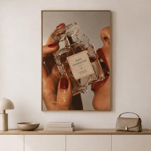 May include: A rectangular wall art print featuring a clear glass perfume bottle with a label that reads MAIN CHARACTER CONFIDENCE IN A BOTTLE. The bottle is held by a hand with dark red nail polish, positioned near a face with red lipstick.