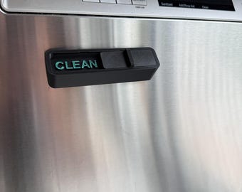 Puede incluir: Un imán negro para lavavajillas con la palabra "CLEAN" en letras turquesas. El imán es rectangular con una cubierta deslizante. El imán está adherido a un lavavajillas de acero inoxidable.
