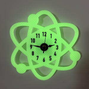 May include: A luminous green atom-shaped wall clock. The clock face has black numerals and hands, with a central black clock mechanism. The outer frame of the clock is a bright, neon green, and the clock is mounted on a light grey wall.