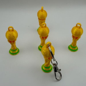 May include: Four yellow trophy-shaped keychains with green circular bases and a small loop at the top. One keychain features a silver metal lobster clasp attached to the loop. The trophies have a textured surface and a rounded top section.